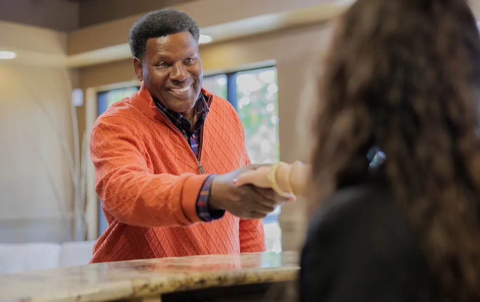A smiling man in an orange quarter-zip sweater shaking hands with a dental receptionist over a marble countertop in a modern office lobby