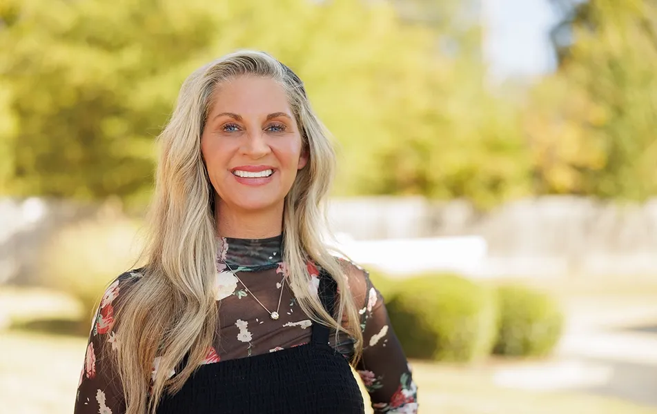 A portrait of a smiling woman with long blonde hair and bright teeth standing outdoors against a soft-focus background of trees and sunshine.