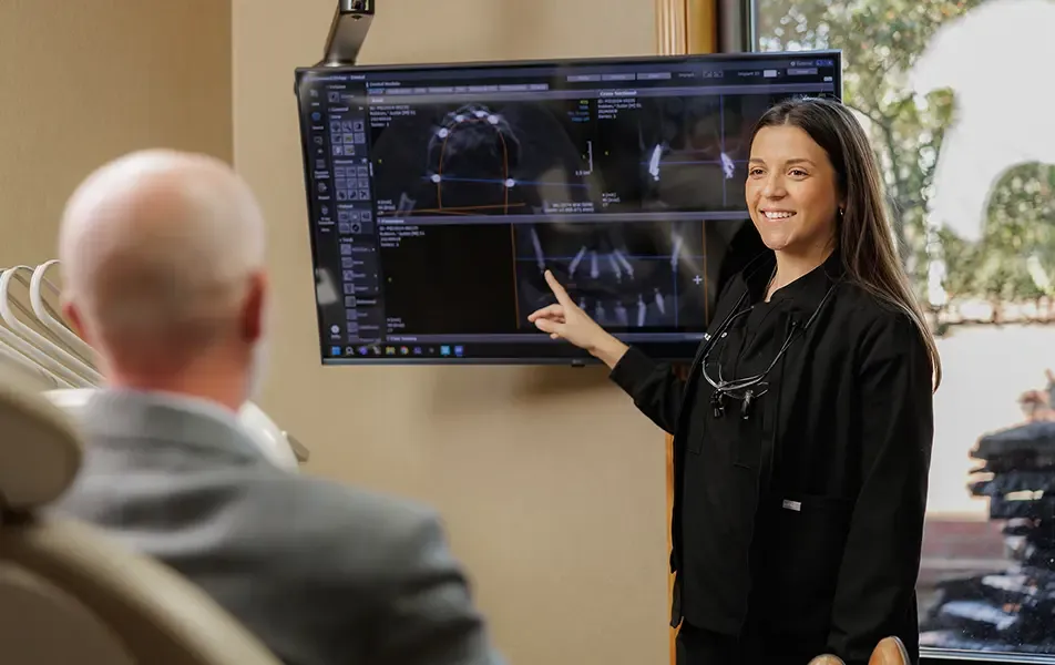 A female dental professional in black scrubs smiling and pointing to a large monitor displaying dental X-rays and 3D scans while a male patient looks on from the dental chair