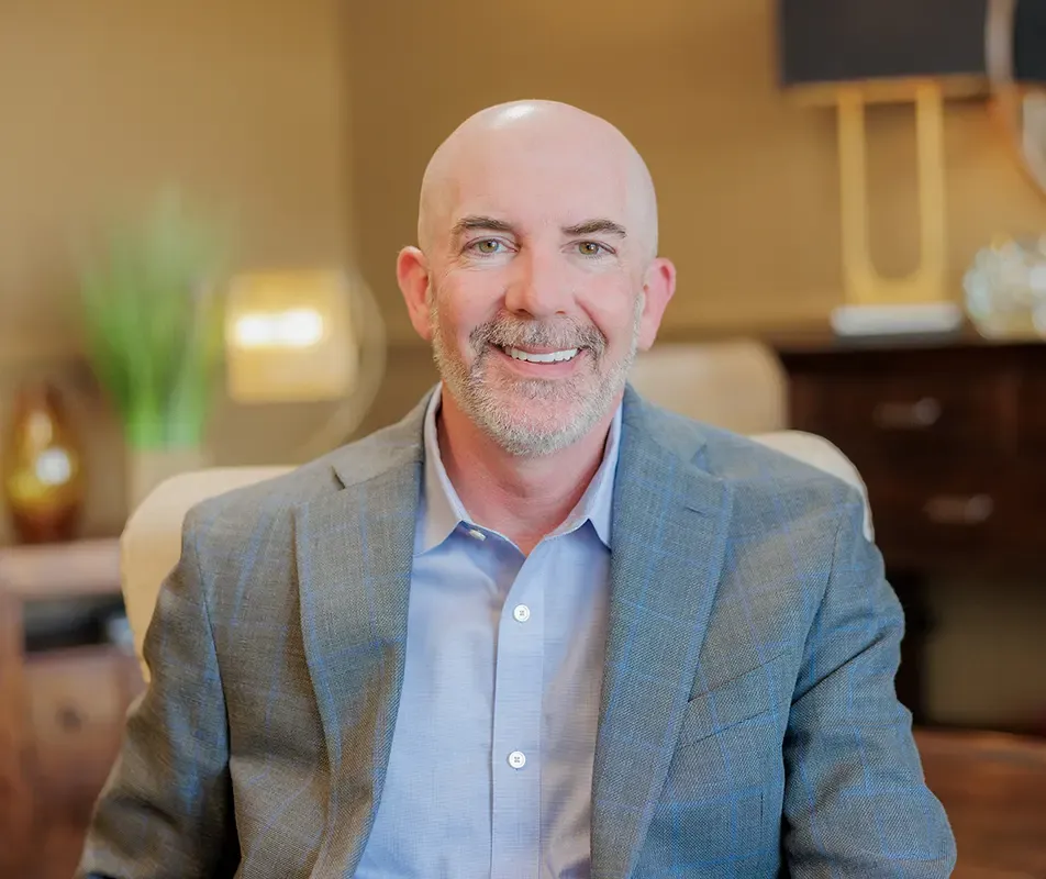 A close-up portrait of a smiling, middle-aged man with a graying beard and a bald head, wearing a blue button-down shirt and a grey plaid blazer